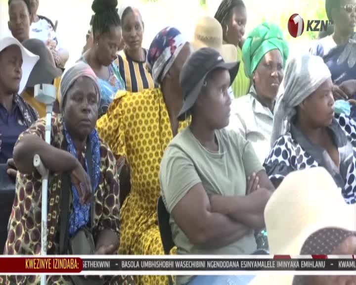 A group of women, some in colourful headscarves, sit attentively. One woman, leaning on a walking stick, looks towards something out of frame. A group of women, some in colourful headscarves, sit attentively. One woman, leaning on a walking stick, looks towards something out of frame.