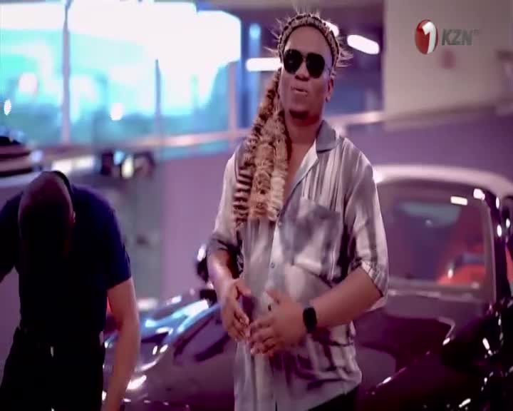 A man in a patterned shirt and sunglasses stands confidently, while another man in a dark shirt bends over beside him. The 1KZN TV logo is visible in the background, suggesting this is a broadcast from South Africa. A man in a patterned shirt and sunglasses stands confidently, while another man in a dark shirt bends over beside him. The 1KZN TV logo is visible in the background, suggesting this is a broadcast from South Africa.