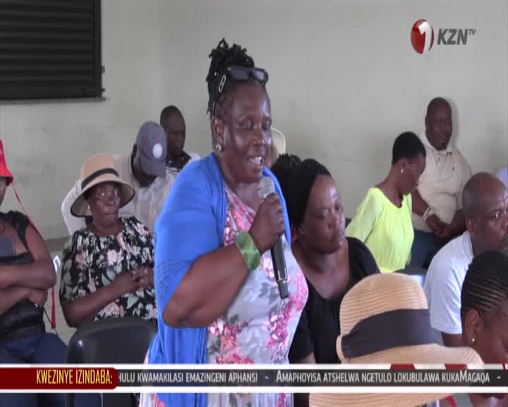A woman in a blue cardigan speaks into a microphone, her arm adorned with green bangles. She's addressing a gathering in what appears to be a community hall, with other attendees seated behind her. A woman in a blue cardigan speaks into a microphone, her arm adorned with green bangles. She's addressing a gathering in what appears to be a community hall, with other attendees seated behind her.