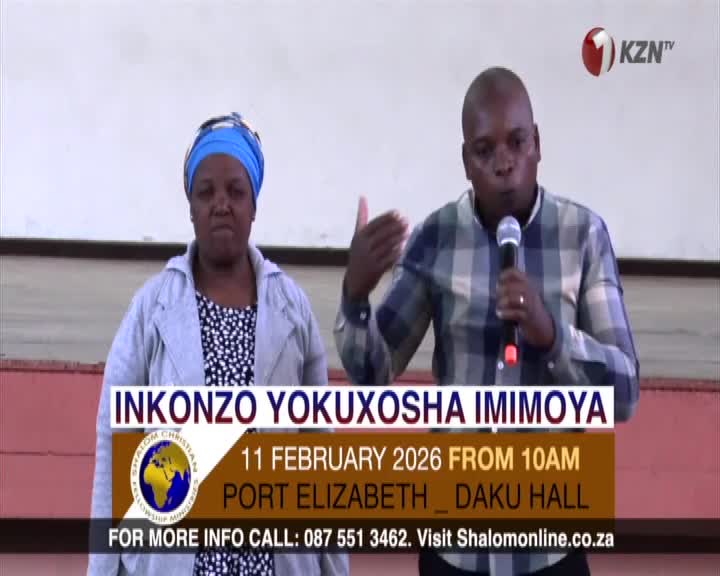 A man speaks into a microphone, his hand gesturing as if emphasizing a point. Beside him, a woman stands attentively, her head covered by a blue scarf. A man speaks into a microphone, his hand gesturing as if emphasizing a point. Beside him, a woman stands attentively, her head covered by a blue scarf.