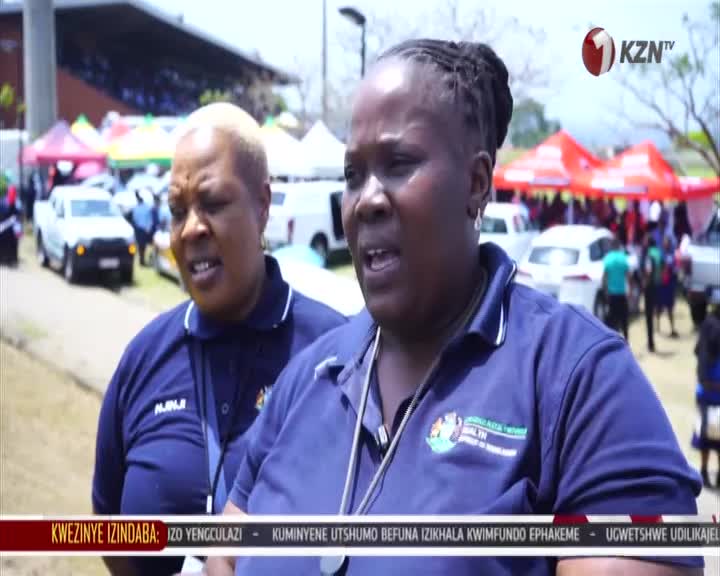 Two women in navy blue shirts stand near a busy event with tents and vehicles in the background. A KZN TV logo appears in the upper right corner. Two women in navy blue shirts stand near a busy event with tents and vehicles in the background. A KZN TV logo appears in the upper right corner.