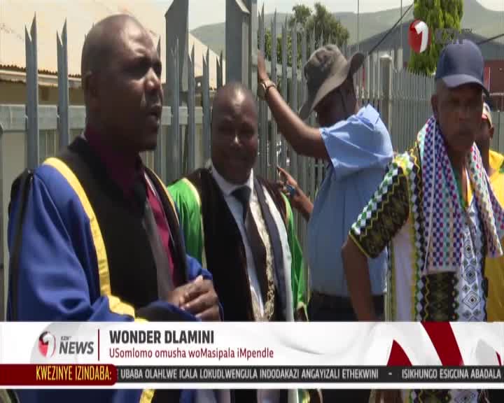 A man in ceremonial robes speaks, his hands gesturing as another man in a blue shirt and hat reaches out. Behind them, a person in a patterned shirt and scarf stands with a blue cap.