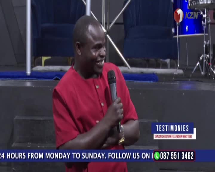 A man in a bright red shirt speaks into a microphone on a stage, his face lit with a broad smile. Behind him, a blue drum with the KZN logo sits ready for a performance.