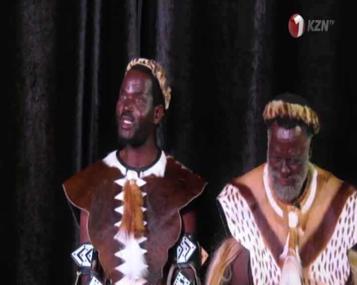 Two men, dressed in traditional Zulu attire, stand before a dark curtain. The man on the left, with a beaded headband, speaks animatedly.