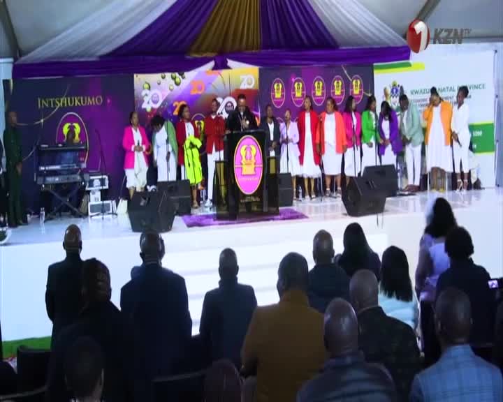 A group of women in bright jackets and white dresses stand on a stage, singing. The KZN TV logo is visible in the corner of the screen.