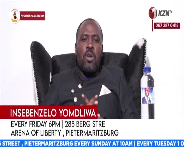 A man in a black suit speaks from a large, ornate chair. A bottle with a picture of him on it sits to his right.