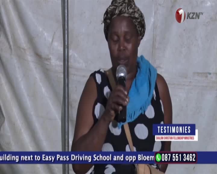 A woman speaks into a microphone, her eyes closed. A blue scarf is draped around her neck.