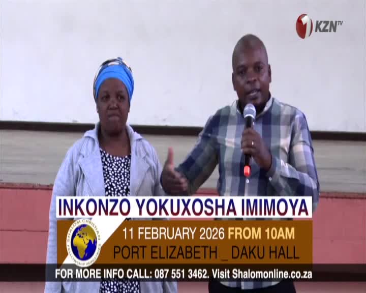 A man in a plaid shirt holds a microphone, speaking to a crowd. Beside him, a woman in a blue headscarf listens intently.