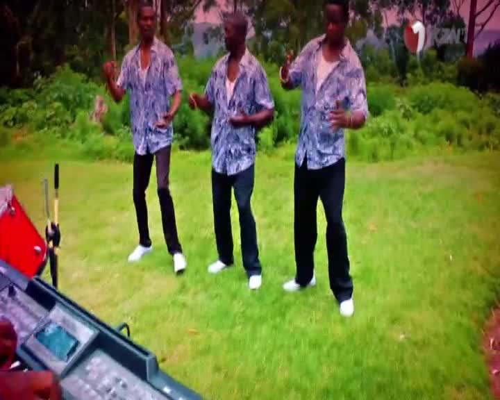 Three men in patterned shirts and black trousers are dancing on the grass. A keyboard and a red drum are set up nearby, ready for a performance.