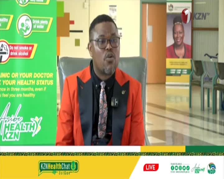 A man in a bright orange suit speaks into a microphone. Behind him, a KZNTV logo is visible on a screen.