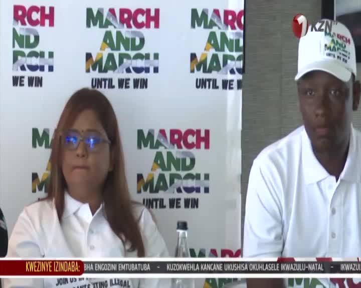 A woman with glasses and a man in a white cap sit behind a table, a banner behind them reads "MARCH AND MARCH UNTIL WE WIN." A 1KZN TV logo is visible in the upper right corner.