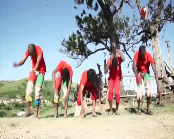 Five men in red shirts and patterned leg coverings bend low in unison, their movements sharp and synchronized. The scene unfolds on a dusty hillside, a familiar backdrop for many performances broadcast by 1KZN TV across South Africa.