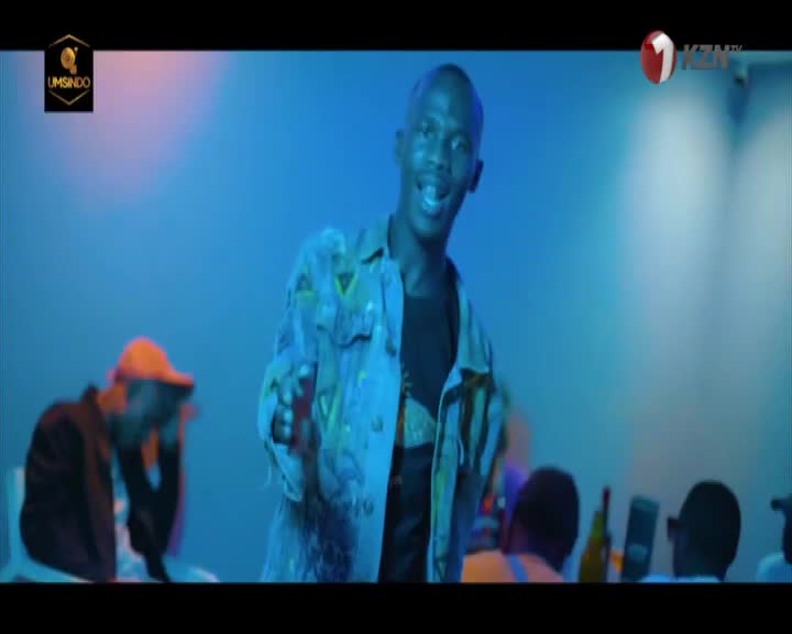 A young man in a patterned jacket gestures forward, his mouth open as if speaking. Behind him, a person in an orange cap leans forward, and other figures are visible in the blue-lit space.