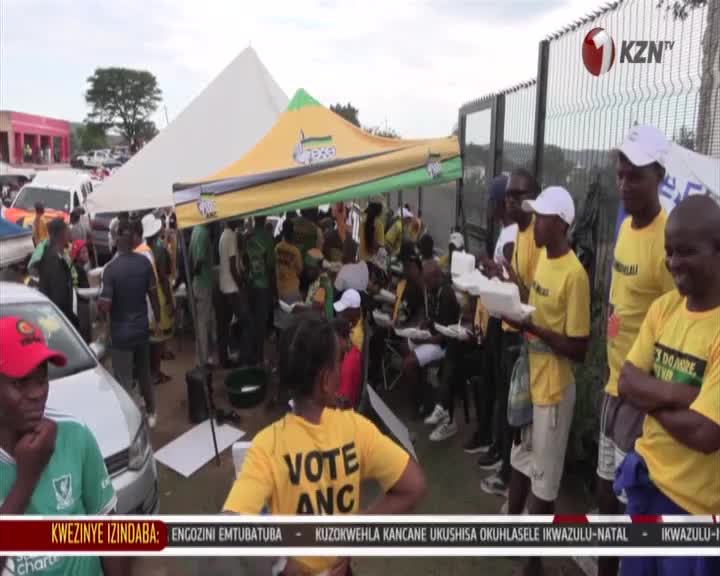 A crowd gathers under yellow and green tents, many wearing ANC t-shirts. People are being served food from white containers.