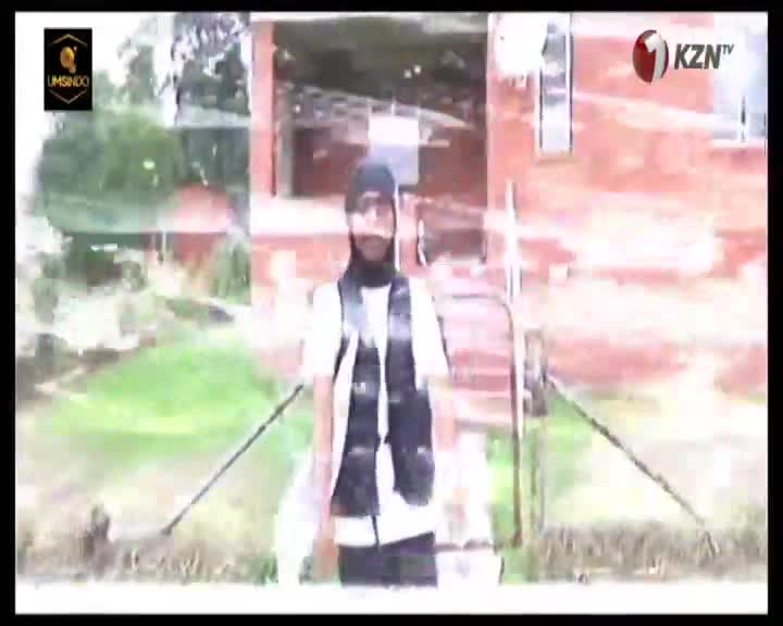 A young man stands on a grassy slope in front of a brick building. He wears a black headscarf, a white t-shirt, and a dark vest.