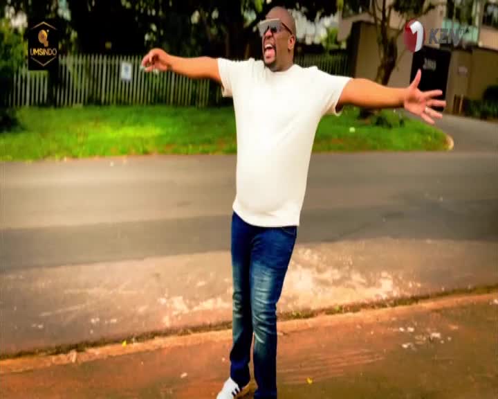 A man in a white t-shirt and blue jeans throws his arms wide open, his mouth agape in a shout. The 1KZN TV logo is visible in the background, suggesting this is a broadcast from KwaZulu-Natal, South Africa. A man in a white t-shirt and blue jeans throws his arms wide open, his mouth agape in a shout. The 1KZN TV logo is visible in the background, suggesting this is a broadcast from KwaZulu-Natal, South Africa.