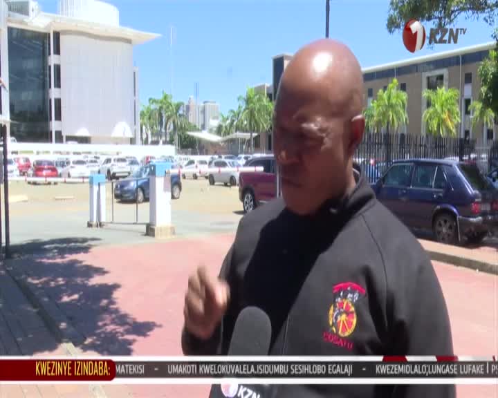 A man in a black jacket with a COSATU emblem speaks into a microphone. Cars are parked in the background of a sunny day in South Africa. A man in a black jacket with a COSATU emblem speaks into a microphone. Cars are parked in the background of a sunny day in South Africa.