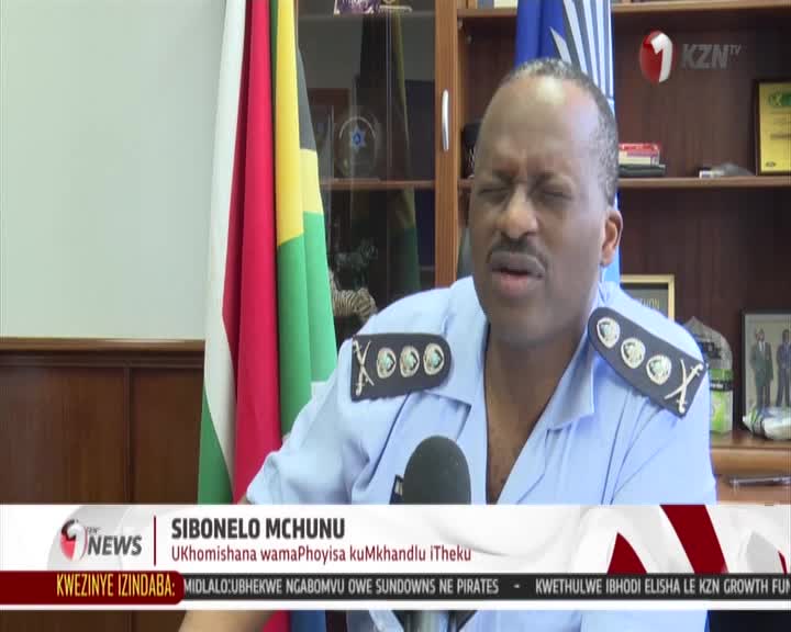 A man in a light blue uniform with epaulets is speaking into a microphone, his eyes closed. Behind him, the South African flag hangs next to another flag, and a KZN TV logo is visible. A man in a light blue uniform with epaulets is speaking into a microphone, his eyes closed. Behind him, the South African flag hangs next to another flag, and a KZN TV logo is visible.