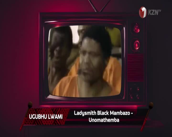 On the screen, a group of men in orange shirts are captured in a moment of performance. The 1KZN TV logo is visible in the corner, identifying the broadcast. On the screen, a group of men in orange shirts are captured in a moment of performance. The 1KZN TV logo is visible in the corner, identifying the broadcast.