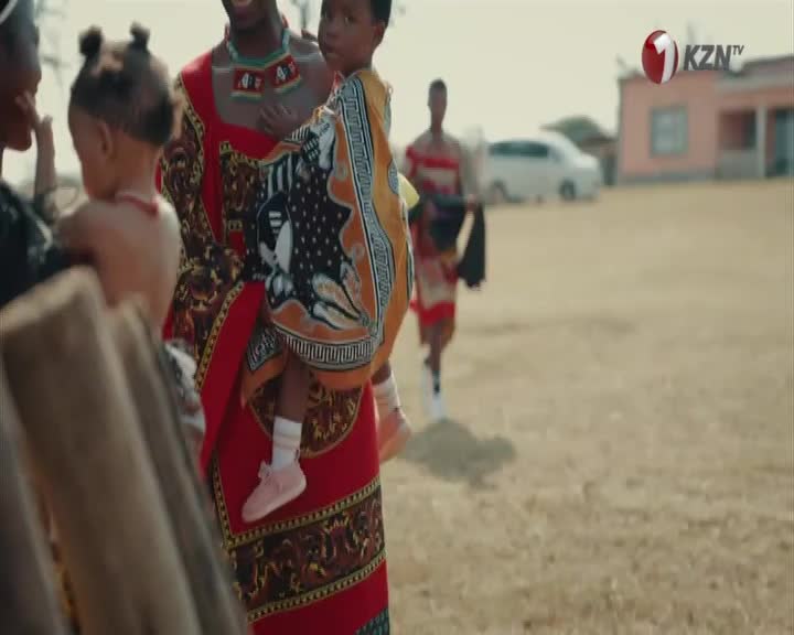 A woman in a vibrant red and gold patterned dress holds a child, her arm wrapped around the little one's back. Another child, with hair styled in two buns, stands close by, their gaze directed away from the camera. A woman in a vibrant red and gold patterned dress holds a child, her arm wrapped around the little one's back. Another child, with hair styled in two buns, stands close by, their gaze directed away from the camera.