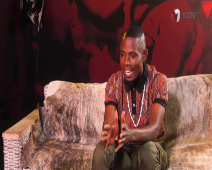 A young man with a sharp haircut, wearing a patterned shirt and a beaded necklace, gestures with his hands as he speaks. He's seated on a rustic couch covered in animal hide, with the 1KZN TV logo visible in the background.