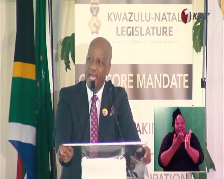 A man in a suit speaks at a podium, the South African flag behind him. A woman signs alongside him, her hands moving with clear intent.
