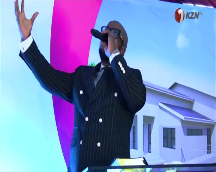 A man in a pinstripe suit holds a microphone to his lips, his left arm raised in a gesture of praise. Behind him, a KZN TV logo is visible against a backdrop of a stylized house.