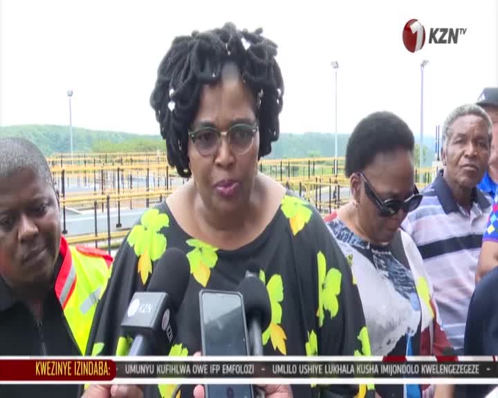 A woman with dreadlocks, wearing glasses and a black dress with yellow flowers, speaks into microphones in front of a KZN TV logo. Behind her, a group of people stand near some yellow industrial structures. A woman with dreadlocks, wearing glasses and a black dress with yellow flowers, speaks into microphones in front of a KZN TV logo. Behind her, a group of people stand near some yellow industrial structures.