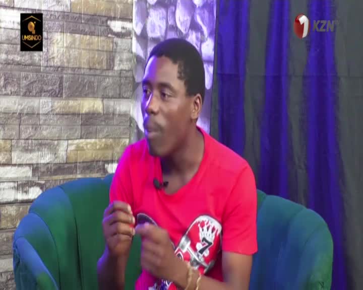 A man in a bright red t-shirt gestures with his hands while speaking, seated on a plush green couch. The KZN television logo is visible in the background. A man in a bright red t-shirt gestures with his hands while speaking, seated on a plush green couch. The KZN television logo is visible in the background.