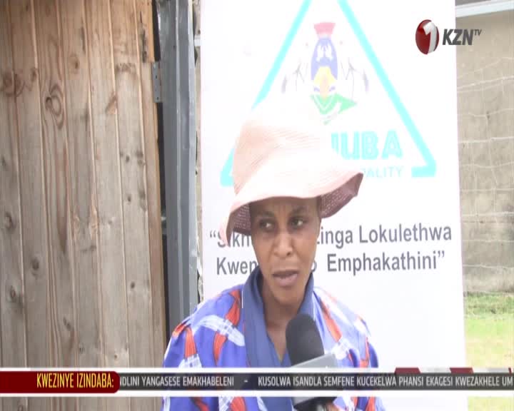A woman in a wide-brimmed hat speaks into a microphone in front of a banner with the 1KZN TV logo. The banner behind her has text in Zulu, likely from a local news report.