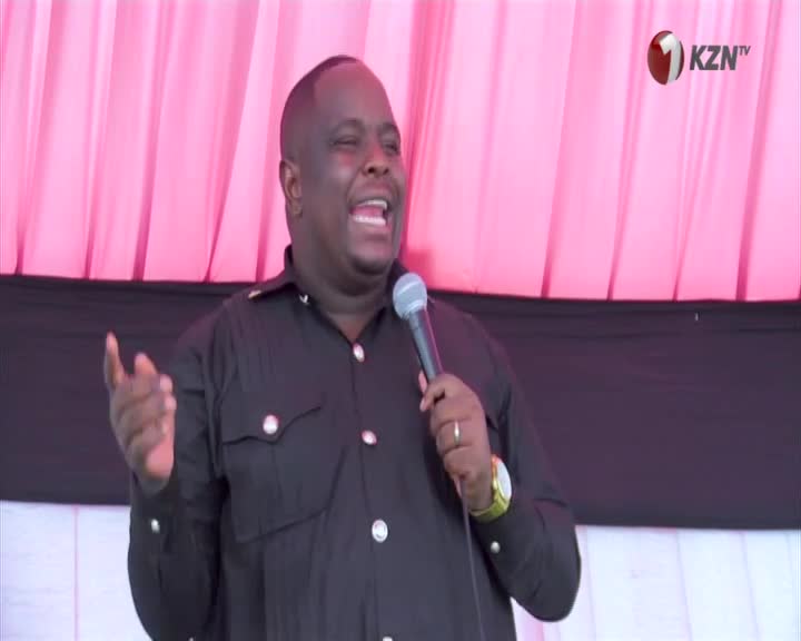 A man in a black shirt speaks into a microphone, his right hand gesturing. The backdrop is a bright pink curtain, with the KZN TV logo visible in the corner.