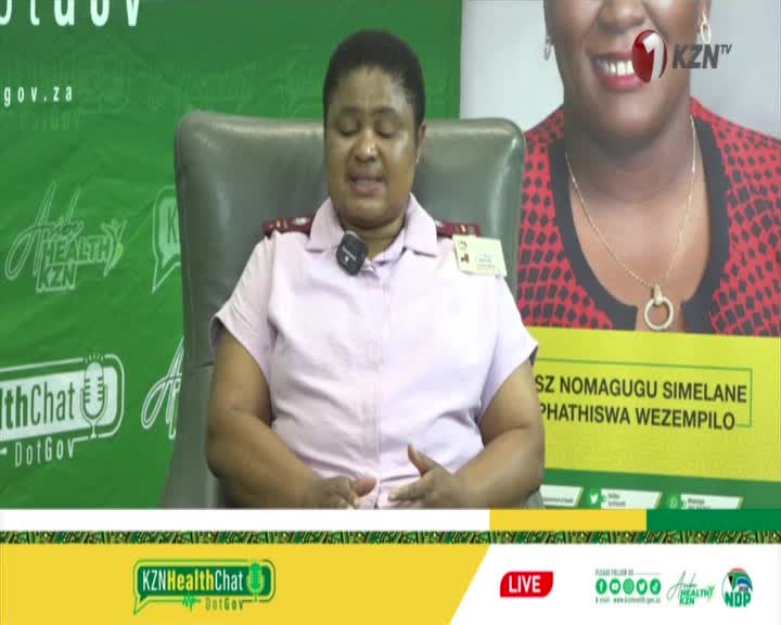 A woman in a light pink uniform speaks into a microphone, her hands clasped in her lap. Behind her, a large KZNTV logo is visible next to a portrait of another woman.