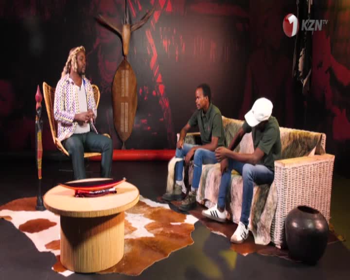 A man with blonde dreadlocks sits in a wicker chair, facing two young men who are seated on a couch. The 1KZN TV logo is visible in the upper right corner of the studio set.