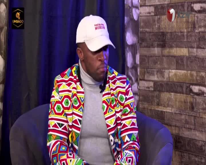 A man in a vibrant, patterned jacket sits on a dark chair, a white cap on his head. The 1KZN TV logo is visible in the background.