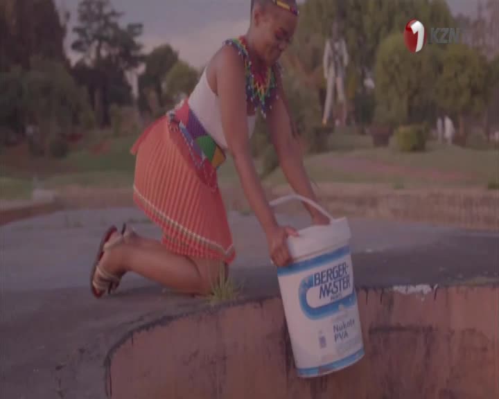 A woman in traditional attire kneels, reaching for a large white bucket. The 1KZN TV logo appears in the corner. A woman in traditional attire kneels, reaching for a large white bucket. The 1KZN TV logo appears in the corner.