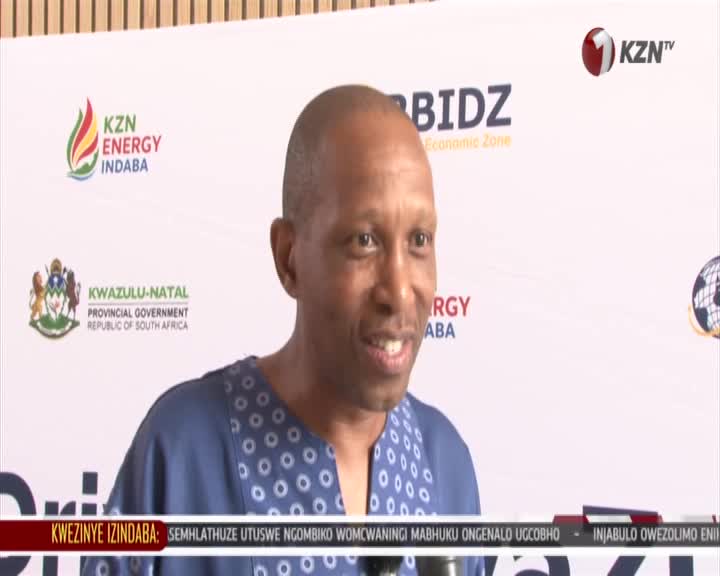 A man in a patterned blue shirt speaks into a microphone. The KZN Energy Indaba logo and the KwaZulu-Natal Provincial Government crest are visible behind him. A man in a patterned blue shirt speaks into a microphone. The KZN Energy Indaba logo and the KwaZulu-Natal Provincial Government crest are visible behind him.