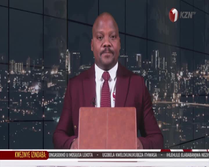 A man in a maroon suit stands behind a wooden podium, a city skyline twinkling behind him. He's presenting the news for 1KZN TV.