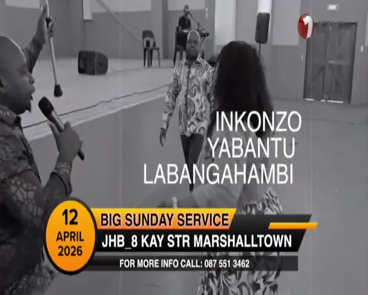 A man in a patterned shirt holds a microphone and a walking stick, speaking with conviction. Another man in a similar shirt walks across the stage, while a keyboardist plays in the background. A man in a patterned shirt holds a microphone and a walking stick, speaking with conviction. Another man in a similar shirt walks across the stage, while a keyboardist plays in the background.