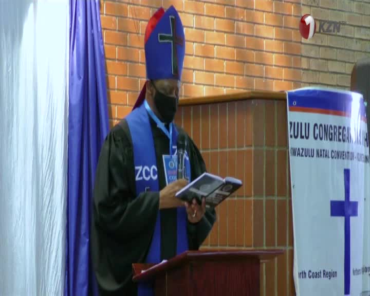 A man in a bishop's mitre and robes reads from a book at a podium. A banner behind him announces the "Zulu Congregation KwaZulu Natal Convention." A man in a bishop's mitre and robes reads from a book at a podium. A banner behind him announces the "Zulu Congregation KwaZulu Natal Convention."