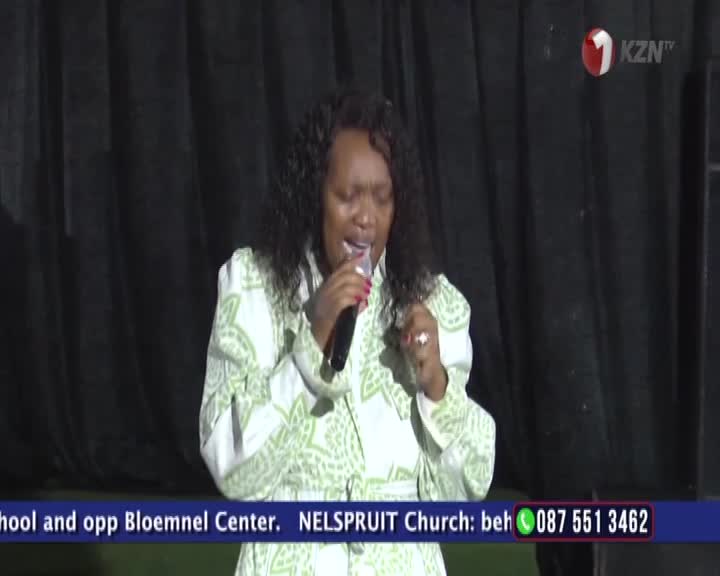 A woman in a green and white patterned dress sings into a microphone, her eyes closed in passion. The 1KZN TV logo is visible in the corner, with text below indicating a church service in Nelspruit. A woman in a green and white patterned dress sings into a microphone, her eyes closed in passion. The 1KZN TV logo is visible in the corner, with text below indicating a church service in Nelspruit.