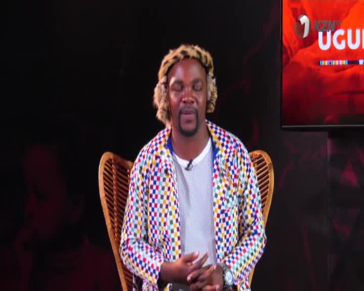 A man wearing a colourful checkered jacket and a leopard-print hat sits in a woven chair, his hands clasped. On a screen behind him, the 1KZN TV logo and "UGULI" are visible. A man wearing a colourful checkered jacket and a leopard-print hat sits in a woven chair, his hands clasped. On a screen behind him, the 1KZN TV logo and "UGULI" are visible.
