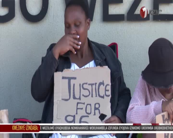 A woman clutches a cardboard sign that reads "Justice For." Another person sits beside her, their face obscured by a hat. The KZN TV logo is visible in the background. A woman clutches a cardboard sign that reads "Justice For." Another person sits beside her, their face obscured by a hat. The KZN TV logo is visible in the background.