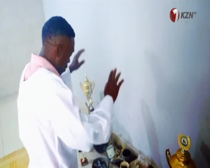A man in a white coat with a pink collar stands with his hands raised, facing a shelf of trophies. The KZN TV logo appears in the upper right corner.