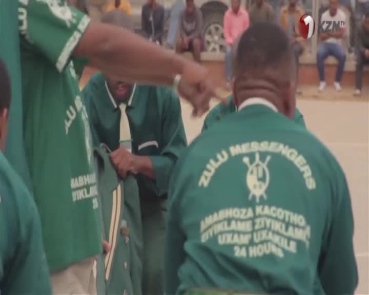A man in a green uniform, emblazoned with "ZULU MESSENGERS," is being handed a jacket. Another man, also in uniform, points towards him. The 1KZN TV logo is visible in the background. A man in a green uniform, emblazoned with "ZULU MESSENGERS," is being handed a jacket. Another man, also in uniform, points towards him. The 1KZN TV logo is visible in the background.