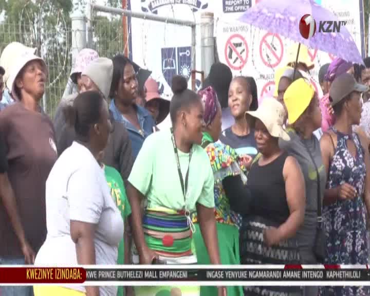 A crowd of people, some wearing hats, gather outside a building. A purple umbrella shields some from the sun while a KZN TV logo is visible. A crowd of people, some wearing hats, gather outside a building. A purple umbrella shields some from the sun while a KZN TV logo is visible.