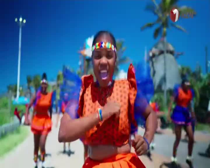 A group of women in vibrant orange and blue traditional attire are dancing energetically along a sun-drenched promenade. Their movements are sharp and rhythmic, a lively display of culture against the bright South African sky, likely a scene for 1KZN TV. A group of women in vibrant orange and blue traditional attire are dancing energetically along a sun-drenched promenade. Their movements are sharp and rhythmic, a lively display of culture against the bright South African sky, likely a scene for 1KZN TV.