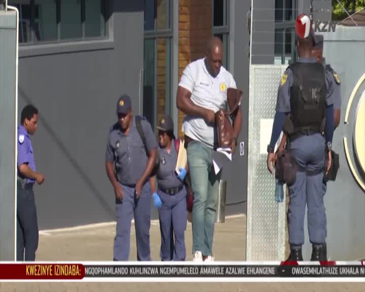 A man in a white polo shirt is rummaging through a brown bag as police officers in uniform stand nearby. A KZN TV crew member, wearing a vest with the station's logo, stands guard at the gate. A man in a white polo shirt is rummaging through a brown bag as police officers in uniform stand nearby. A KZN TV crew member, wearing a vest with the station's logo, stands guard at the gate.