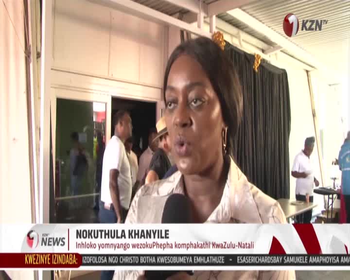 A woman is speaking into a microphone, her mouth open mid-sentence. Behind her, people mill about near an open doorway, some in white shirts. A woman is speaking into a microphone, her mouth open mid-sentence. Behind her, people mill about near an open doorway, some in white shirts.