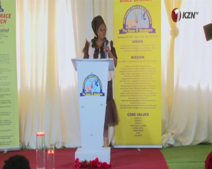A woman in a headscarf speaks from behind a podium in front of a large yellow banner detailing the vision and mission of Abundant Grace Bible Church. The 1KZN TV logo is visible in the upper right corner. A woman in a headscarf speaks from behind a podium in front of a large yellow banner detailing the vision and mission of Abundant Grace Bible Church. The 1KZN TV logo is visible in the upper right corner.