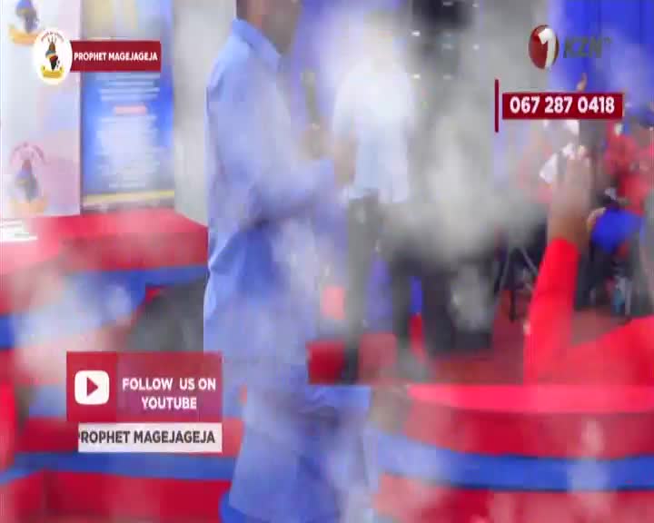 A man in a blue shirt speaks from a stage in South Africa, while a crowd in red shirts watches. The 1KZN TV logo appears on screen with a phone number. A man in a blue shirt speaks from a stage in South Africa, while a crowd in red shirts watches. The 1KZN TV logo appears on screen with a phone number.